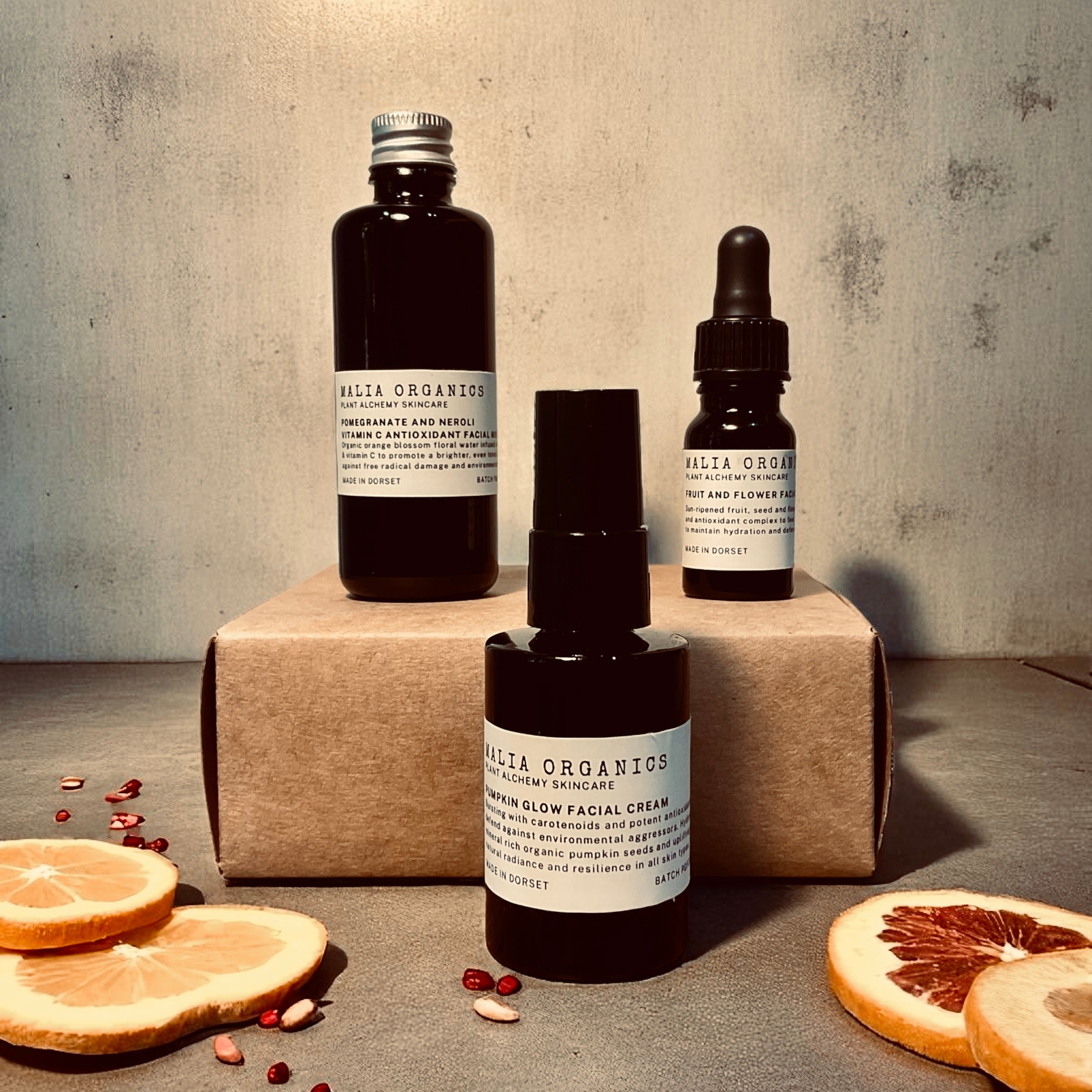 Three bottles of Malia Organics skincare products on a textured surface with dried oranges.